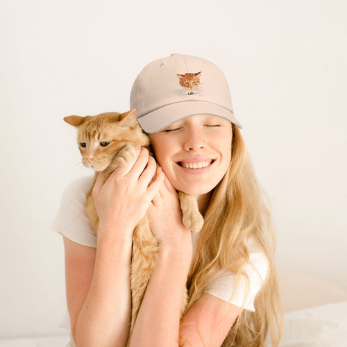 A woman with long blonde hair smiling while holding an orange tabby cat, wearing a light beige baseball cap with an embroidered cat face on the front.